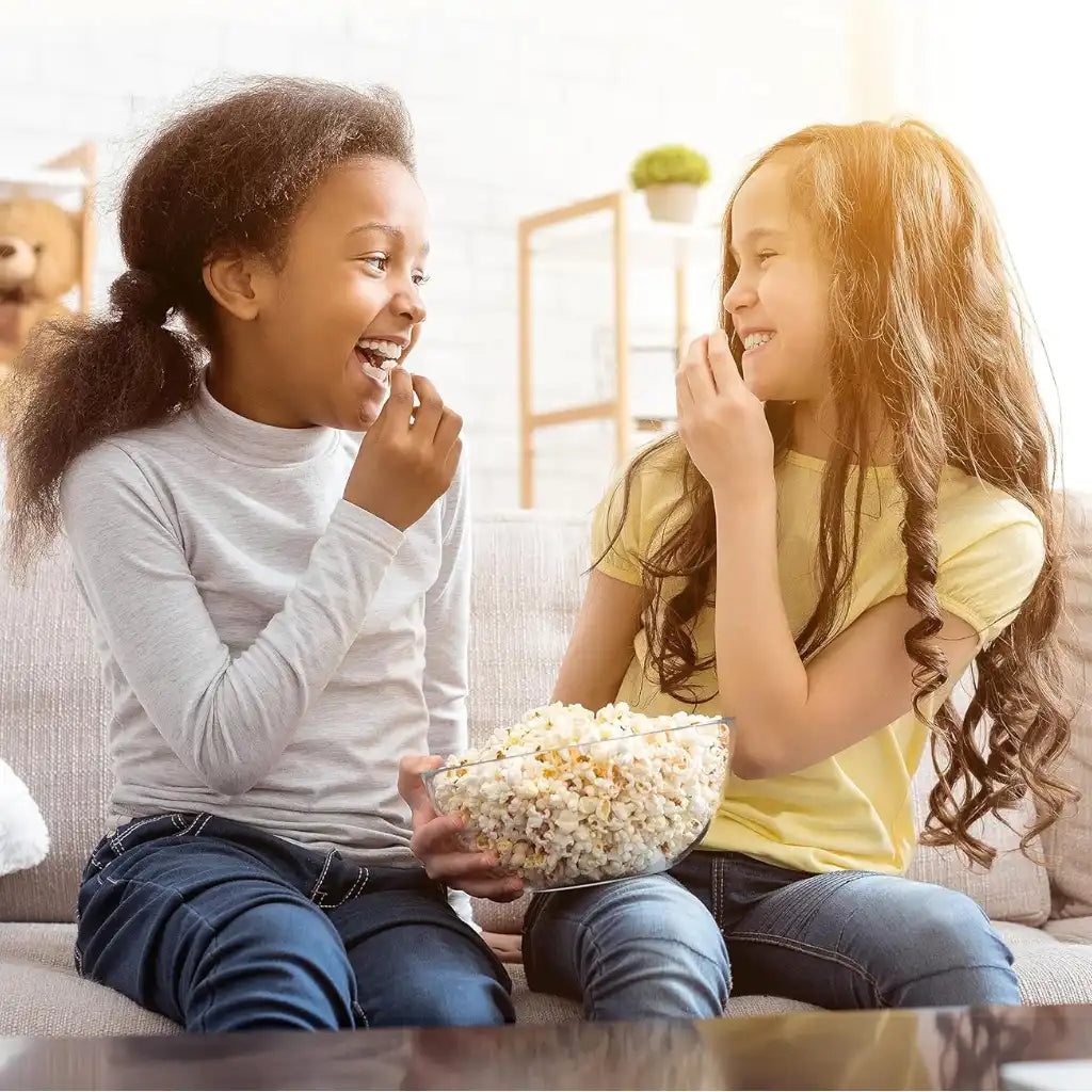Two young girls enjoying popcorn together on a couch.